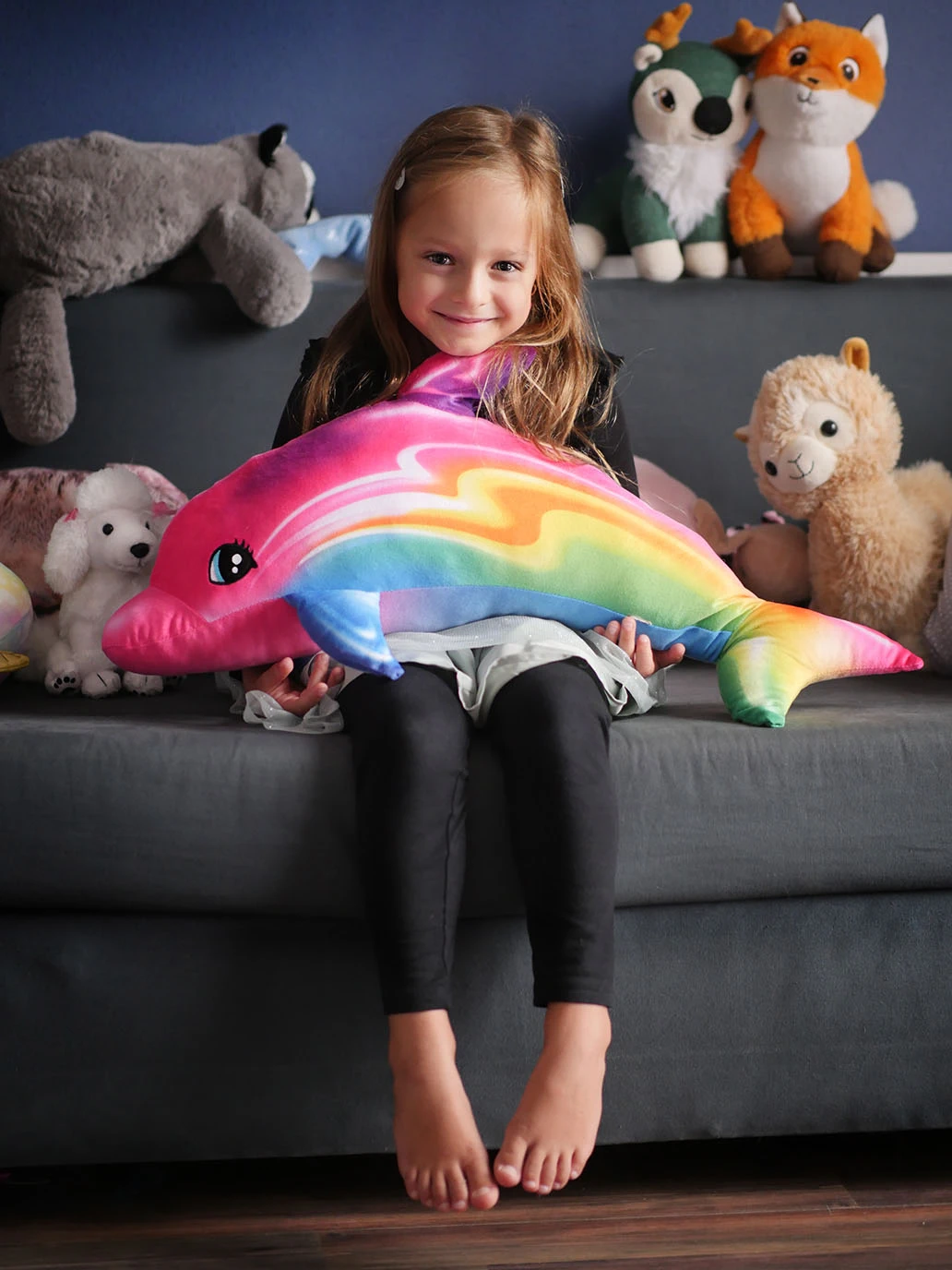 Ein junges Mädchen sitzt auf einem Sofa und lächelt in die Kamera. Sie hält einen riesigen, bunten Delfin in den Händen und ist umrungen von lustigen Stofftieren.