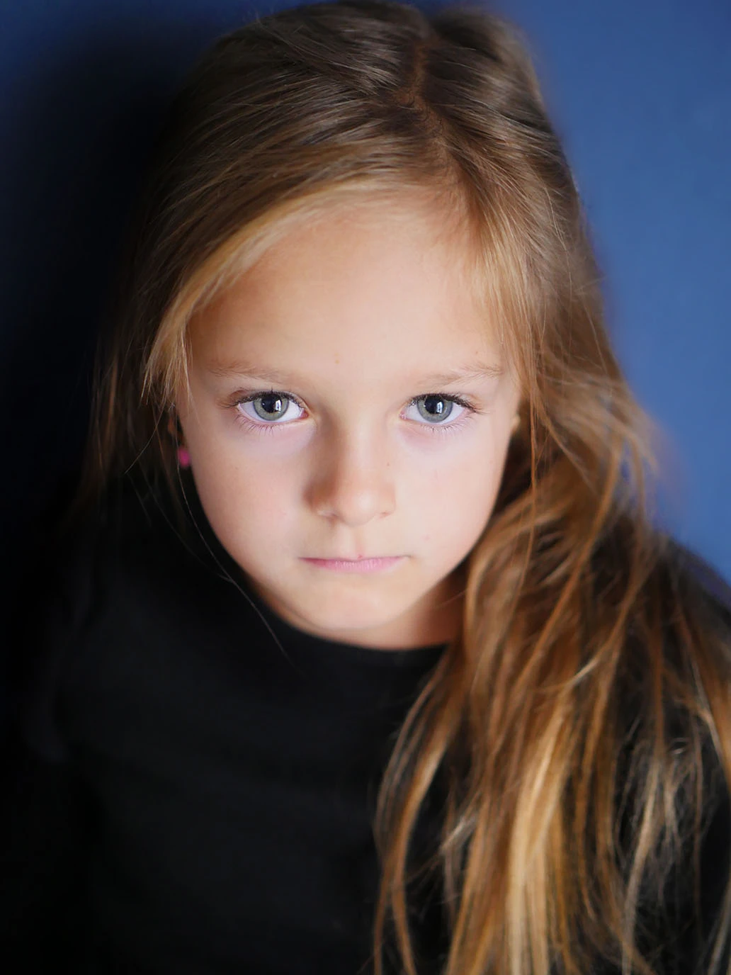 Vor einer blauen Wand blickt ein junges Mädchen geheimnisvoll und mit tiefem Blick in die Kamera. Portraitaufnahme.