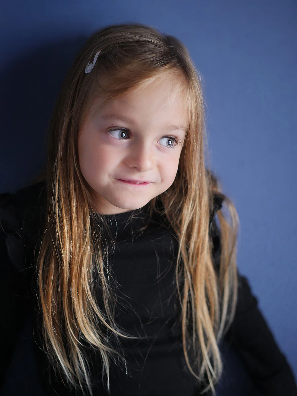 Vor einer Blauen Wand blickt ein Mädchen neugierig und witzig zur Seite. Portraitaufnahme.
