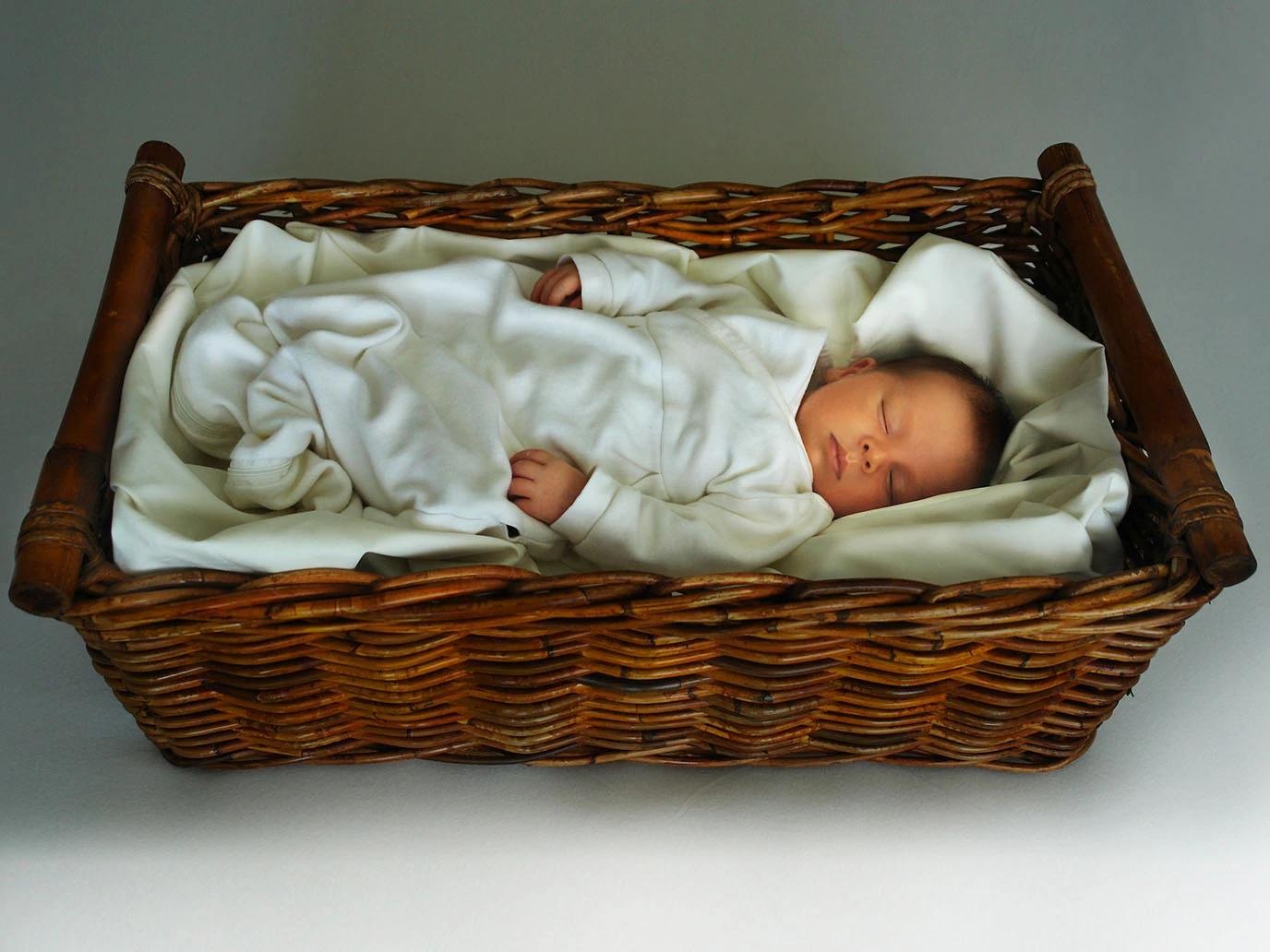 Ein Baby schläft ruhig und geborgen in einem Holz-Korb, zugedeckt von einer weißen Decke.