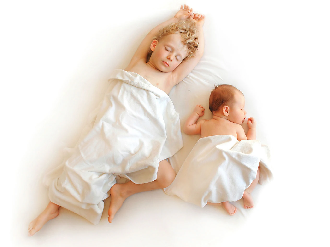 Zwei Geschwister, der eine noch ein Baby, der andere schon ein kleiner Junge, schlafen entspannt nebeneinander in einem weißen Bett.