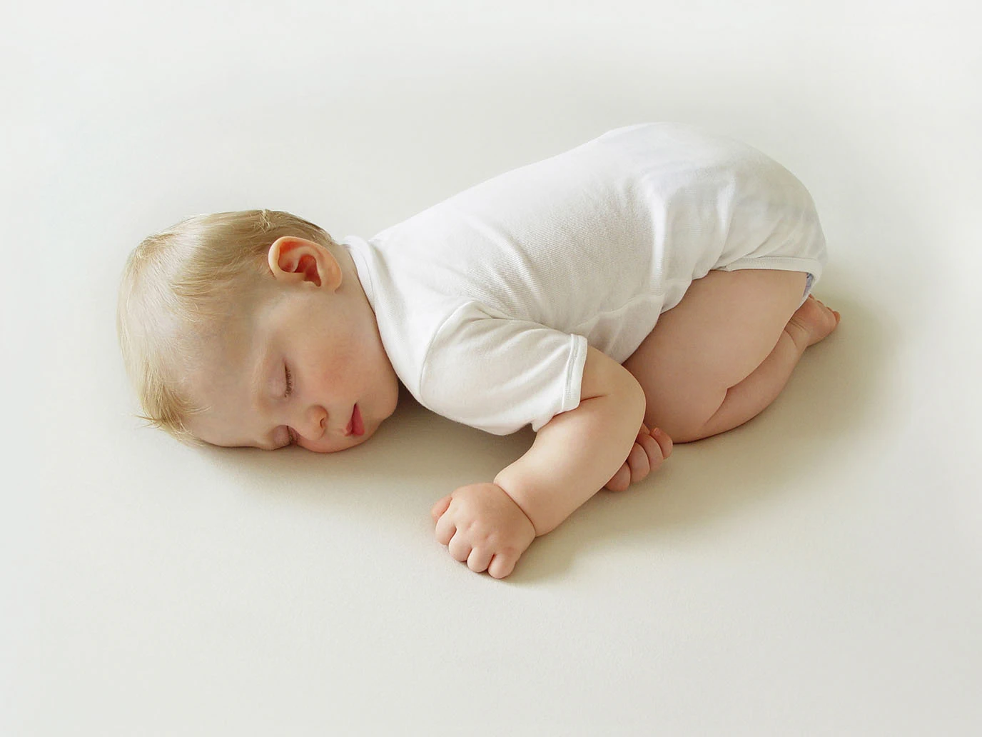  Ein Baby liegt schlafend auf einem weißen Bett. Um das Baby herum ist nichts außer weißer Fläche, es scheint fast wie auf einer Wolke.